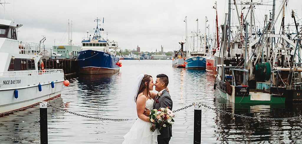 two people embracing in the middle of a traditional new england fishing dock. its a grey cloudy day, they are smiling. one is in a wedding dress. the other in a suit.
